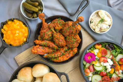 A spread of fried chicken, macaroni and cheese, salad, pickles, and rolls on a wooden board.