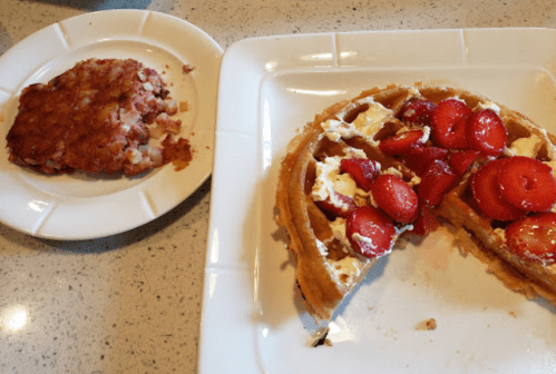 A plate with a sliced waffle topped with strawberries and whipped cream, alongside a serving of hash browns.