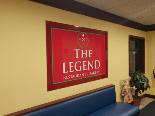 Sign for "The Legend Restaurant & Bakery" on a yellow wall, with a blue bench and a decorative doll nearby.