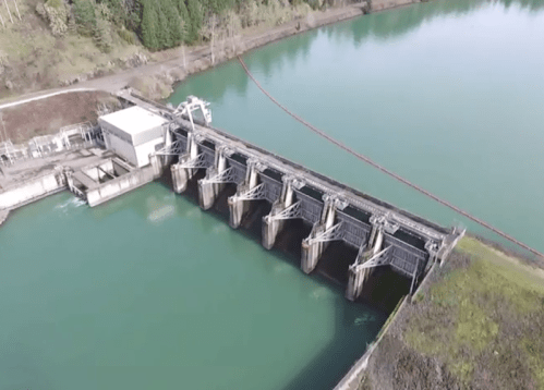 Aerial view of a dam with multiple gates, surrounded by water and trees.