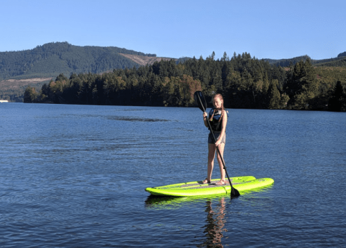 A person stands on a bright green paddleboard in calm water, surrounded by trees and mountains under a clear blue sky.