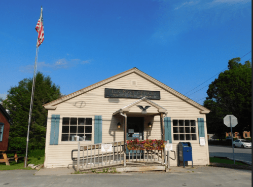 A small U.S. Post Office building with a flag, blue shutters, and flower boxes under a clear blue sky.