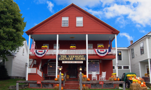 A red country store with white trim, decorated for fall, featuring pumpkins and American flags.