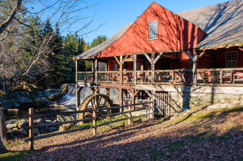 A red barn-style building with a water wheel beside a stream, surrounded by trees and autumn leaves.