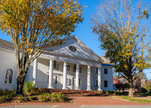 A white building with columns and a circular window, surrounded by trees with autumn leaves. Clear blue sky above.