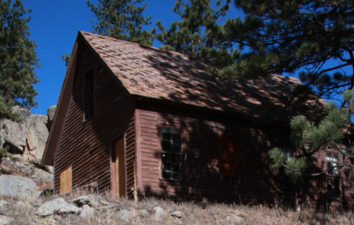 A weathered wooden cabin nestled among trees and rocks under a clear blue sky.