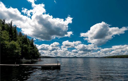 A serene lake scene with a wooden dock, surrounded by lush trees and a bright blue sky filled with fluffy clouds.