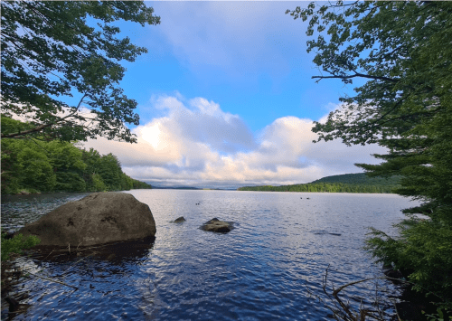 A serene lake surrounded by trees under a blue sky with clouds, featuring a large rock in the foreground.