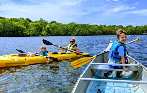 A woman and two children kayak on a calm lake surrounded by greenery on a sunny day.