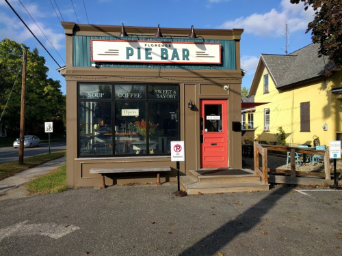 A quaint pie bar with a colorful sign, outdoor seating, and large windows, set on a sunny street.
