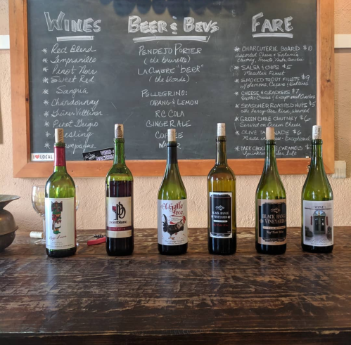 A wooden table with six wine bottles in front of a chalkboard menu listing wines, beers, and food options.