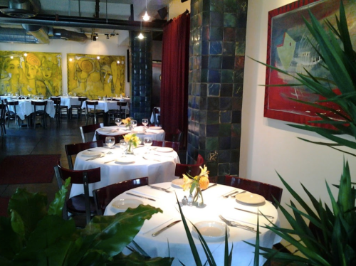 A cozy restaurant interior with white tablecloths, colorful artwork, and plants, creating an inviting atmosphere.