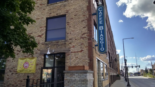 Exterior of Caffe Biaggio, a brick building with a sign, trees, and a clear blue sky in the background.
