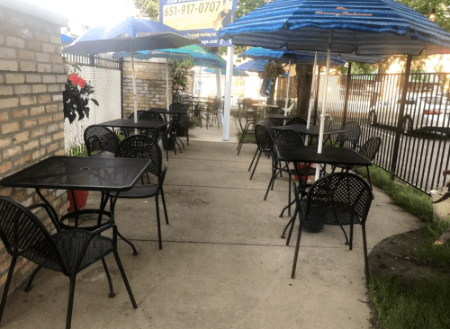 Outdoor dining area with black tables and chairs, shaded by blue umbrellas, along a brick wall.