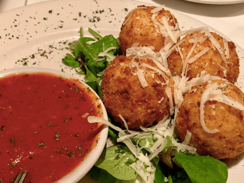Four golden-brown fried balls on a bed of greens, served with marinara sauce and sprinkled with cheese and herbs.