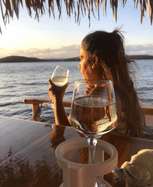 A woman enjoys a drink by the water at sunset, with a glass of wine in the foreground and a scenic view behind her.