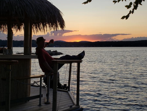 A person relaxes on a dock at sunset, silhouetted against the colorful sky and calm water.