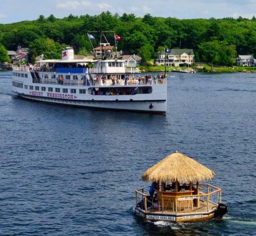 A large white boat with passengers sails on a lake, while a smaller tiki-style boat floats nearby. Green trees line the shore.