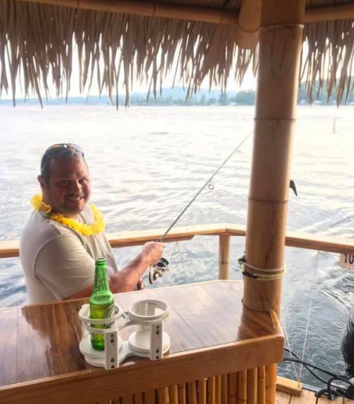 A man wearing a lei smiles while fishing from a bamboo hut by the water, with a drink on the table beside him.
