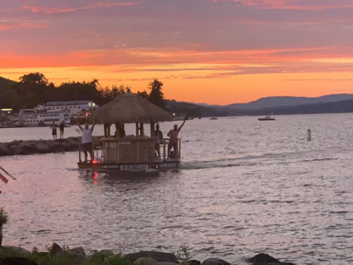 A floating hut on a lake at sunset, with two people waving and boats in the background.