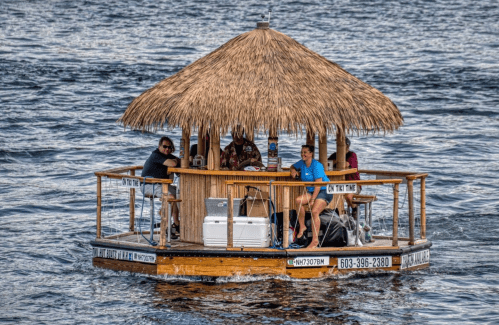 A small, thatched-roof floating bar with people enjoying drinks on a lake.