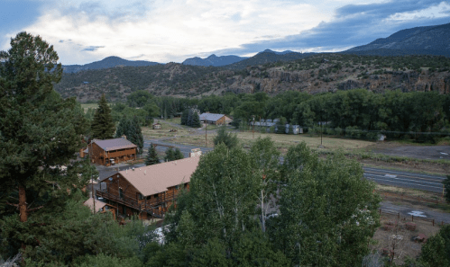 A scenic view of a rural landscape with wooden cabins, trees, and mountains under a cloudy sky.