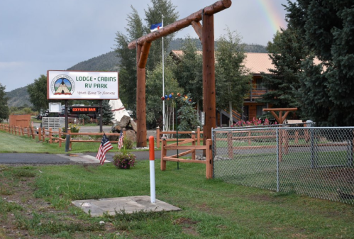 Sign for a lodge and RV park with wooden structures, greenery, and a rainbow in the background.