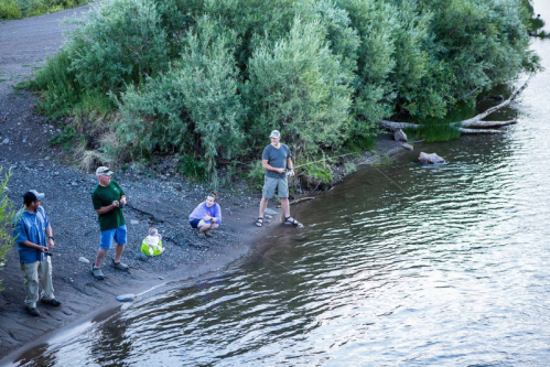 A group of five people fishing by a river, surrounded by greenery, with one child sitting on the shore.