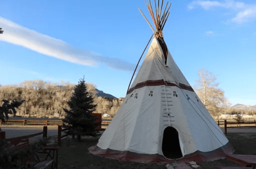 A traditional teepee stands in a scenic outdoor setting with trees and mountains in the background under a clear blue sky.