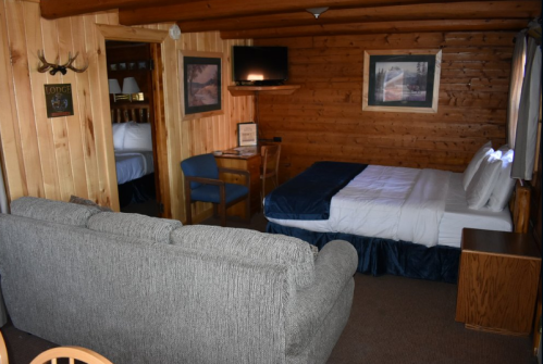 Cozy cabin interior featuring a bed, sofa, desk, and wooden walls, with a TV and framed pictures.