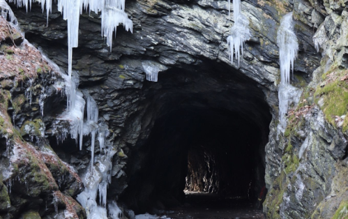 A dark cave entrance framed by icicles and mossy rocks, with a hint of light visible inside.