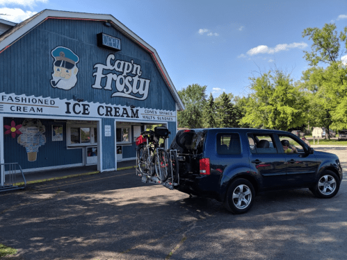 A blue SUV parked outside Cap'n Frosty ice cream shop, with bicycles mounted on the back. Sunny day, trees in the background.