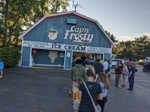 A crowd lines up outside Cap'n Frosty ice cream shop, featuring a blue building with an open sign and ice cream offerings.