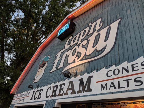 Sign for "Cap'n Frosty" ice cream shop with a blue wooden exterior and neon "OPEN" sign. Cones and malts available.