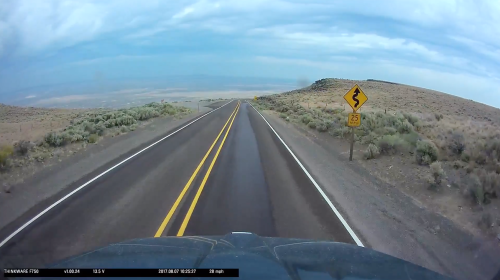 A winding road descends a hill with a warning sign for a 25 mph speed limit ahead. Cloudy sky in the background.