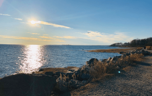 A serene shoreline at sunset, with sunlight reflecting on the water and a rocky path along the coast.