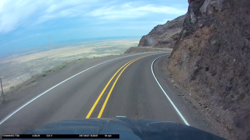 A winding road along a cliff with a vast landscape and cloudy sky in the background.