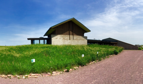 A modern house with a sloped roof, surrounded by tall grass and a gravel driveway under a blue sky.