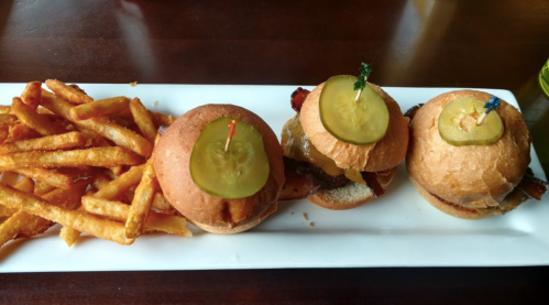 Three mini burgers topped with pickles, served alongside a portion of crispy fries on a white plate.