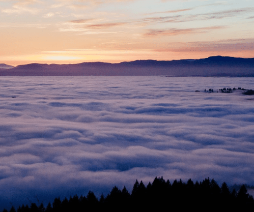 A serene landscape of rolling fog covering the ground, with mountains silhouetted against a colorful sunset sky.