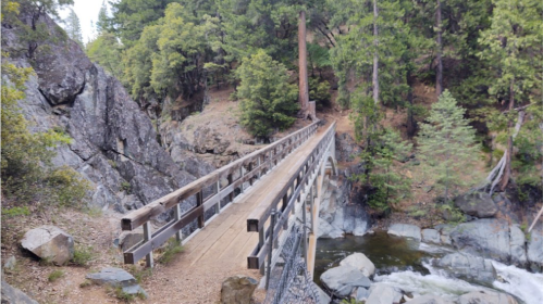 A wooden bridge spans a rocky river, surrounded by lush green trees and rugged terrain.