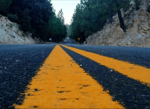 A winding road with yellow double lines, flanked by trees and rocky terrain, under a clear sky.