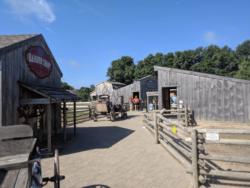 A rustic village scene with wooden buildings, a barber shop sign, and people near a horse-drawn carriage under a clear sky.