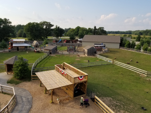 Aerial view of a farm with animal pens, wooden structures, and a playground, surrounded by green fields and trees.
