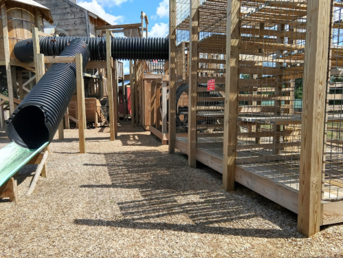Playground structure featuring slides, tunnels, and wooden enclosures, surrounded by gravel and sunlight.