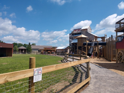 A playground with wooden structures and slides, surrounded by green grass and blue skies with fluffy clouds.