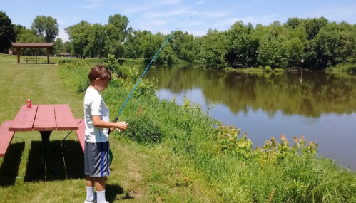 A young boy stands by a calm river, fishing with a rod, while a picnic table is visible nearby.