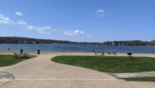 A serene lakeside view with picnic tables, green grass, and a clear blue sky.