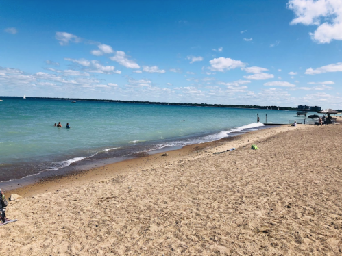 A sandy beach with clear blue water, people swimming, and a few clouds in a bright sky.