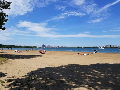 A sandy beach with people relaxing, blue water, and a city skyline in the background under a clear blue sky.